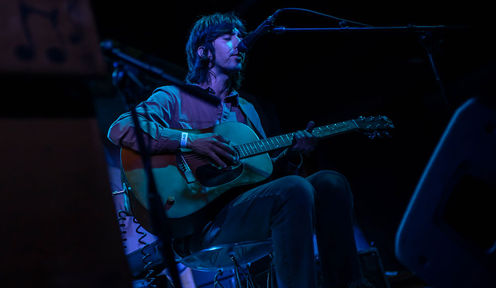 Person playing guitar onstage, dark background with blue highlights