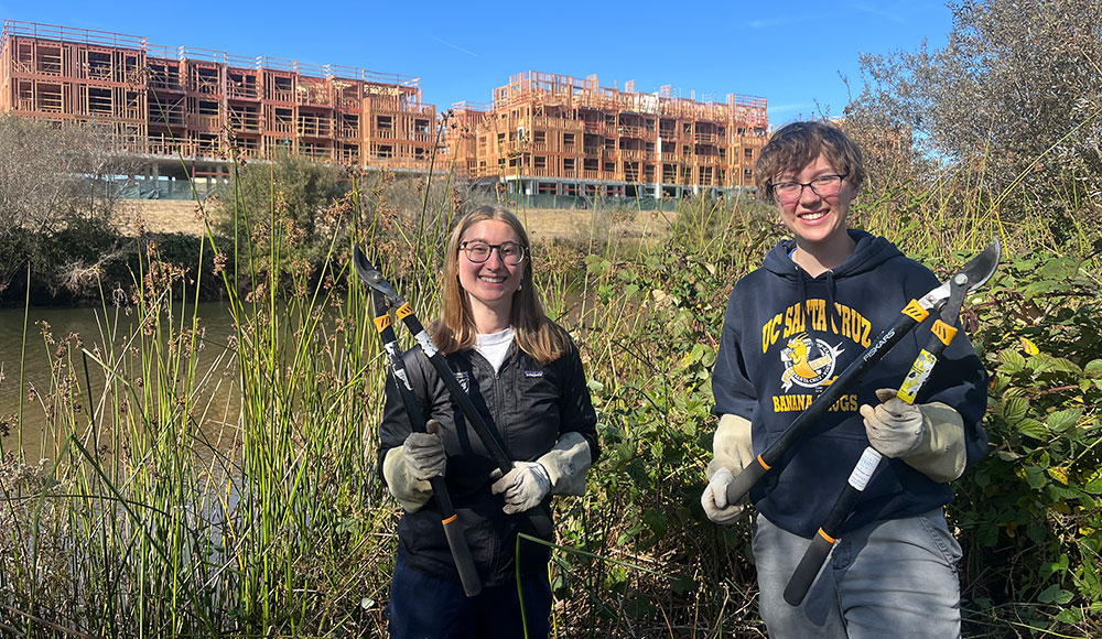 Two women with clippers in front of heavy vegetation; in the background, a river with a construction project above it