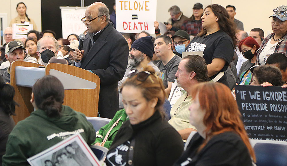 Man speaking at a meeting with people holding up signs