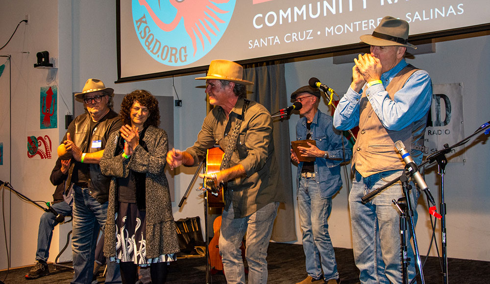 Musicians up on a stage in front of a banner for radio station KSQD